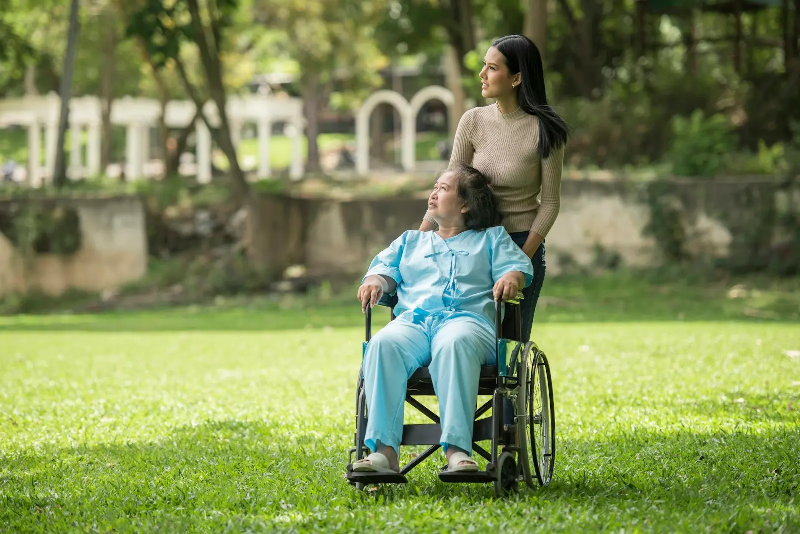 A caregiver walking beside an older woman in a wheelchair on a sunny afternoon